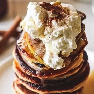 A stack of pancakes topped with whipped cream and a sprinkle of cinnamon sits on a white plate. Syrup drips down the sides of the pancakes. A blurred setting in the background includes cinnamon sticks and a small bowl.