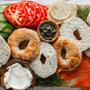 A wooden board with an assortment of bagels, cream cheese, smoked salmon, cucumbers, tomatoes, red onion slices, capers, and a lemon wedge arranged neatly for Mother's Day serving.