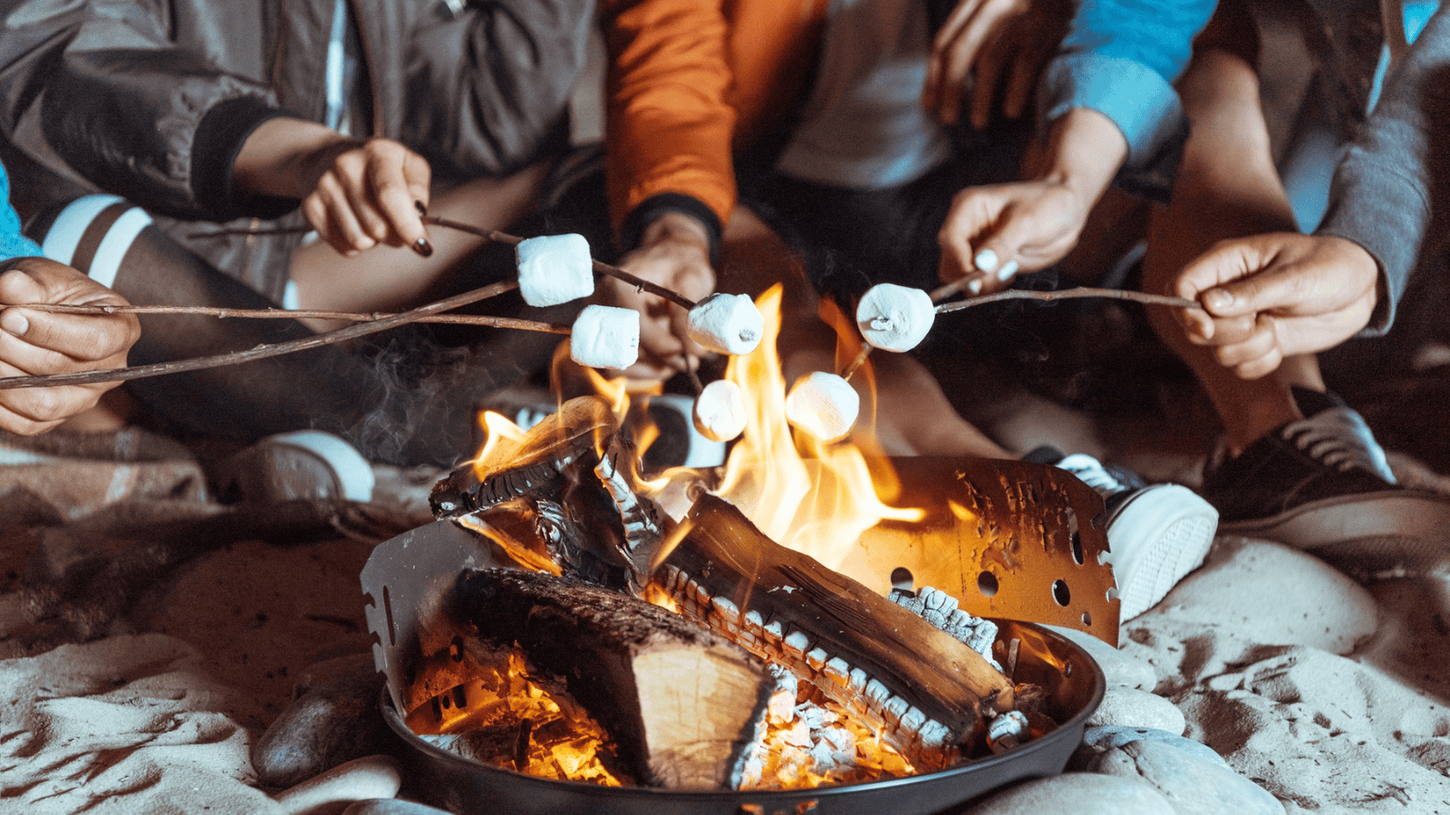 A group of people sit around a small campfire toasting marshmallows on sticks. The fire is contained in a metal container, and the scene is set on a sandy surface. Only the hands of the people are visible.
