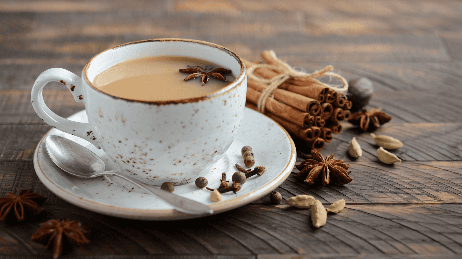 A white cup filled with chai tea sits on a matching saucer with a spoon. The tea is garnished with a star anise. Surrounding the cup are various spices including cinnamon sticks, cardamom pods, star anise, and peppercorns on a wooden table.