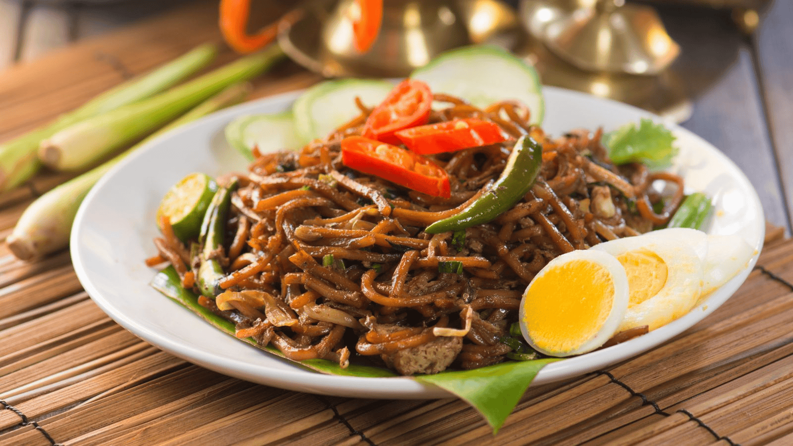 A plate of noodle stir-fry with slices of boiled egg, green chilies, and red chili slices. The dish is garnished with cucumber slices and served on a leaf with bamboo mat and lemongrass stalks visible in the background.