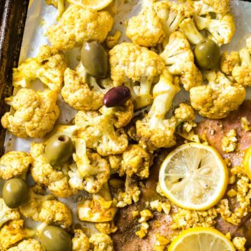 Roasted cauliflower with lemon slices and olives on a baking tray, accompanied by a small bowl of sauce. The dish is presented in a rustic style, featuring vibrant yellow and green colors inspired by recipes