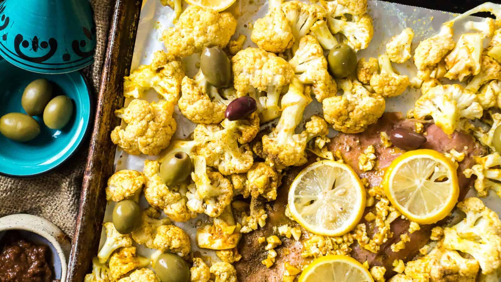 Roasted cauliflower with lemon slices and olives on a baking tray, accompanied by a small bowl of sauce. The dish is presented in a rustic style, featuring vibrant yellow and green colors inspired by recipes