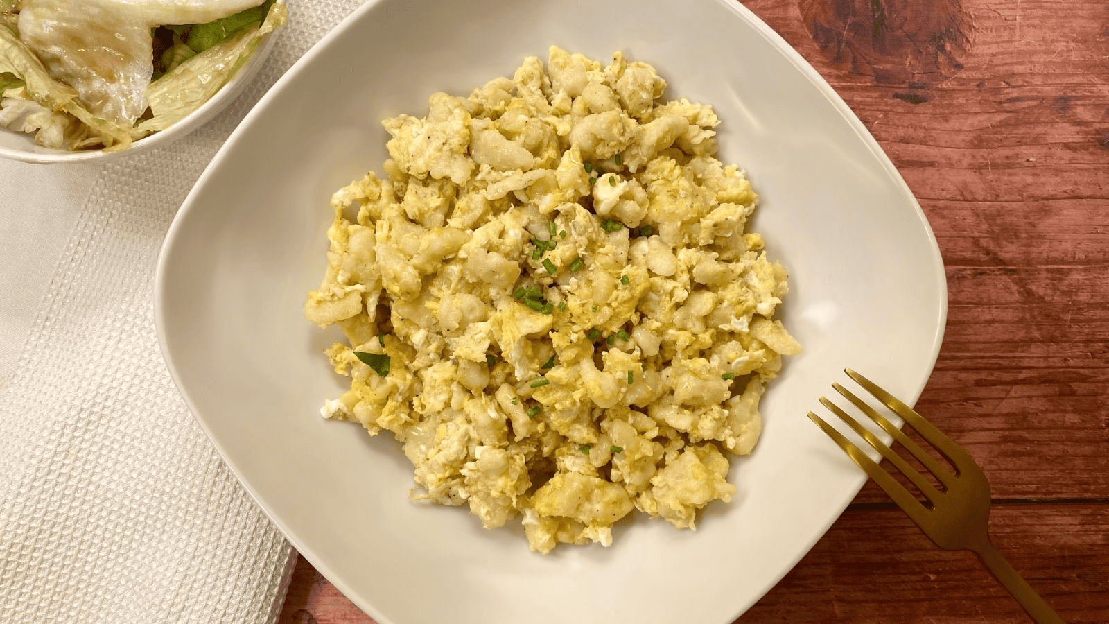 A bowl filled with scrambled eggs and broken pieces of matzo. The dish is topped with small green herbs and placed on a white square-shaped plate. A gold fork is on the right side, and there's a partially visible salad bowl in the top left corner.