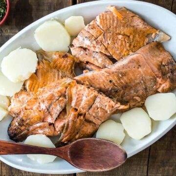 A plate of grilled salmon fillets and boiled potato slices, garnished with herbs, served on a rustic wooden table with a wooden serving spoon and a bowl of sauce on the side.