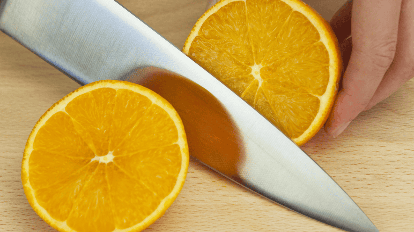 A close-up image of an orange being sliced in half by a knife on a wooden surface. One hand is holding one half of the orange while the other half rests on the surface.