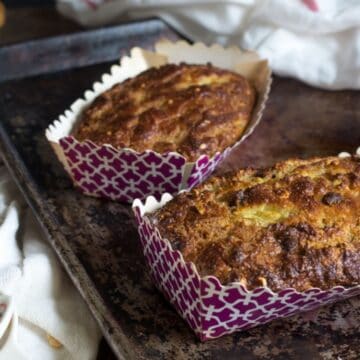 Two freshly baked banana breakfast breads sit in decorative pink and white paper trays on a rustic metal baking sheet. Surrounding the tray are various baking supplies, including chocolate chips, nuts, and a colorful measuring cup, all placed on a pristine white cloth.