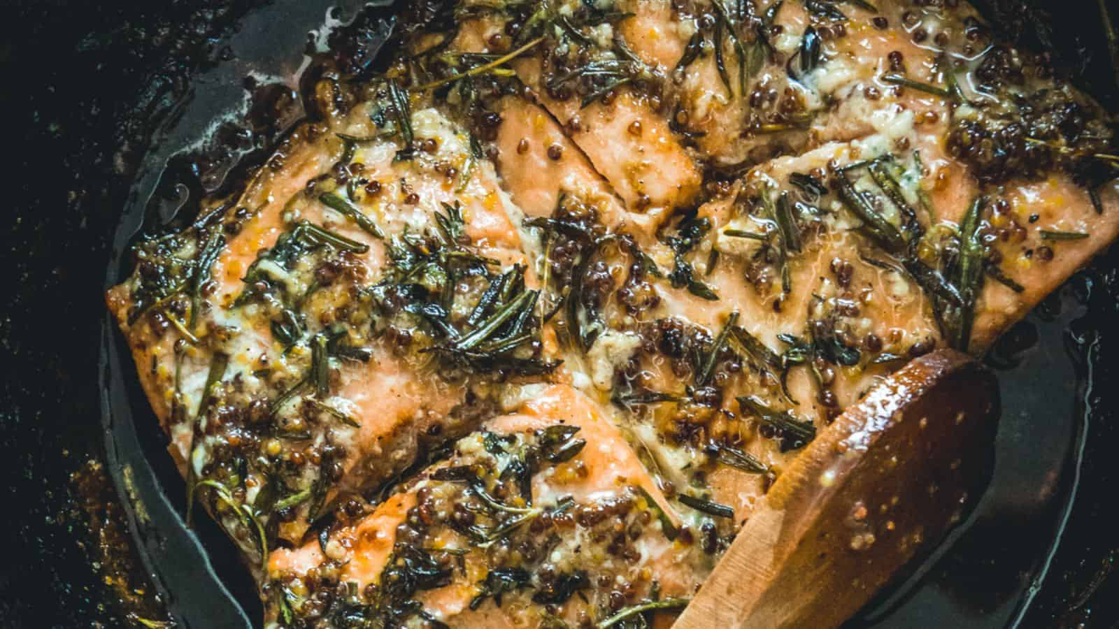 A salmon fillet being cooked in a skillet, garnished with herbs and a glaze, with a wooden spoon stirring the sauce around it.