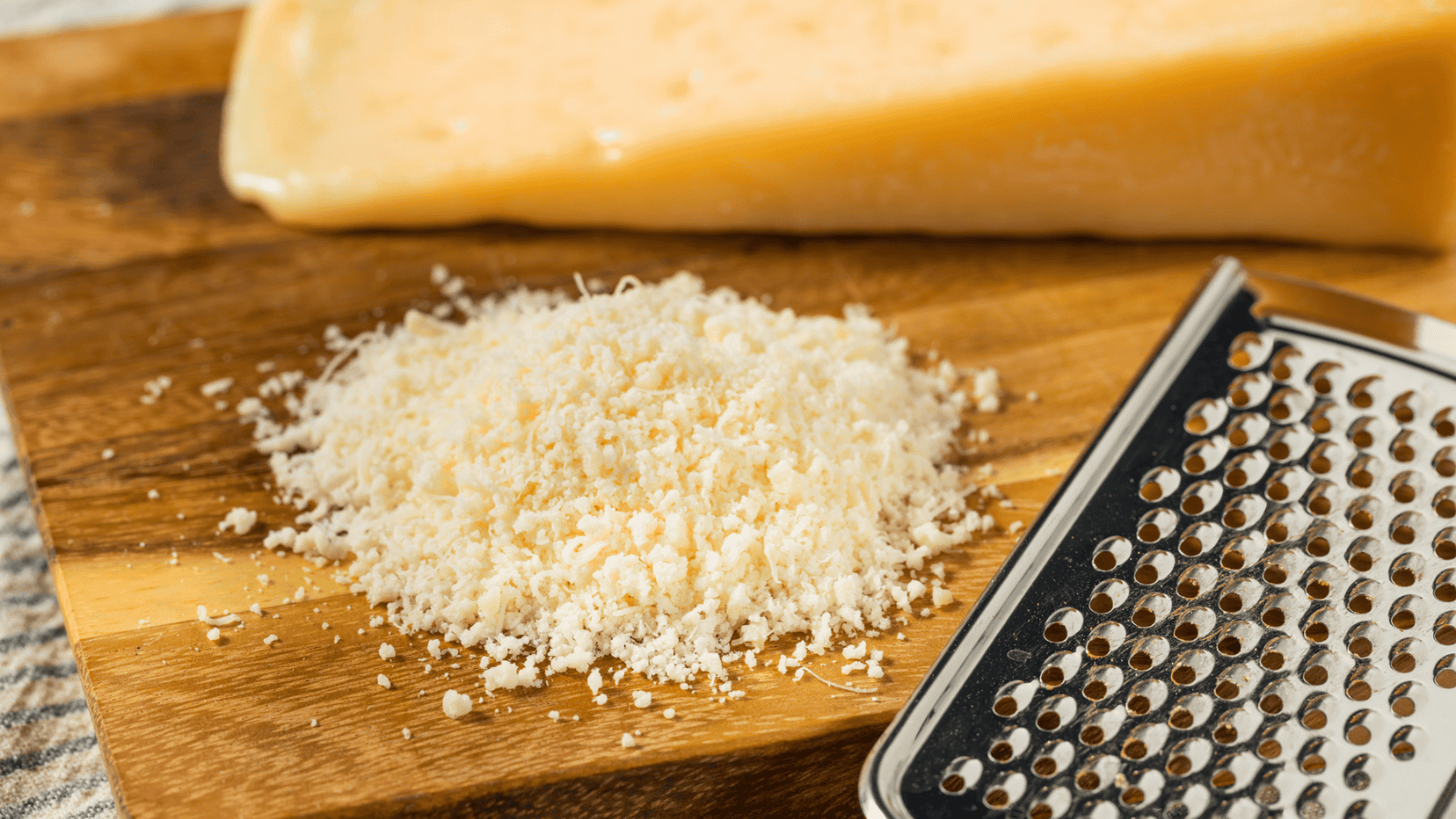 A chunk of Parmesan cheese sits on a wooden cutting board with a pile of grated cheese next to it. A metal grater is positioned to the right of the pile.