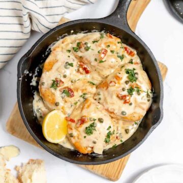 A skillet containing creamy garlic chicken with herbs and red pepper flakes, garnished with parsley and lemon slices. a striped towel and pieces of bread are beside the pan.