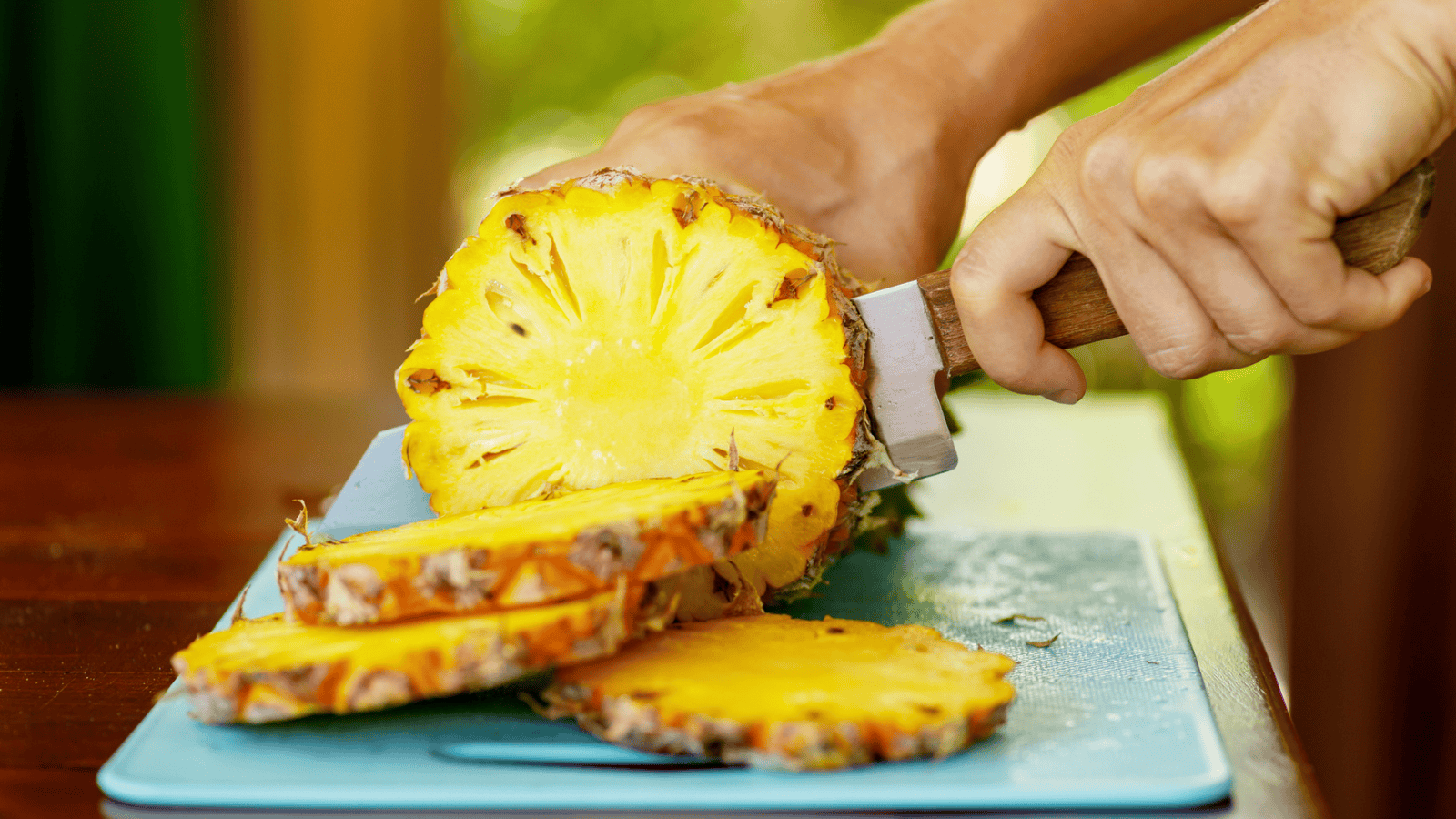 A person slicing a pineapple on a blue cutting board, with two pineapple slices already cut and lying on the board. The background is a mix of green and yellow, suggesting an outdoor setting.