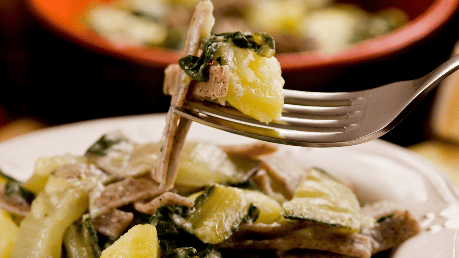 A close-up image of a fork holding a bite of pasta and vegetables, likely potatoes and greens. In the background, more of the same dish is visible in a bowl. The pasta appears to be topped with a light sauce.
