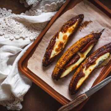 Three baked plantains topped with melted cheese and placed on a parchment-lined baking sheet. A white, crocheted tablecloth partially covers the background, and a knife with a wooden handle rests beside the baking sheet.