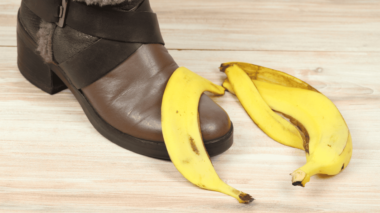 A brown leather boot stepping on a banana peel, with another banana peel nearby, placed on a light wooden floor.