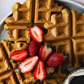 A plate of four golden brown waffles topped with halved strawberries. A small jug of syrup is placed next to the waffles. The plate is on a light-colored surface, with two folded cloth napkins nearby.