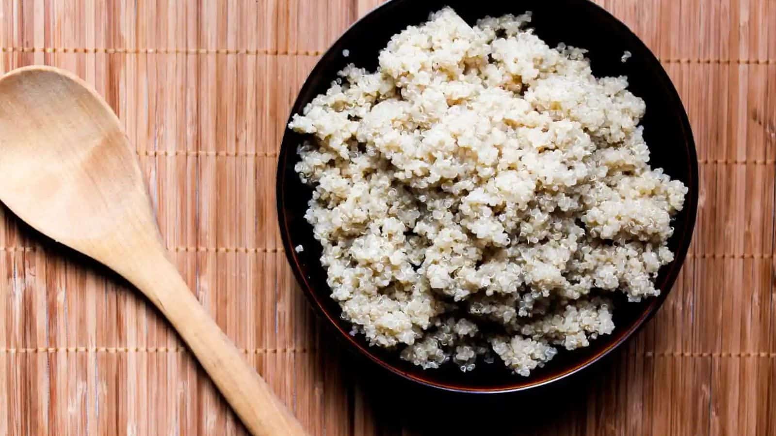 A bowl of cooked quinoa on a bamboo mat with a wooden spoon to the side. The quinoa, part of our Veggie-Powered Proteins series, appears fluffy and is served in a dark, round bowl.