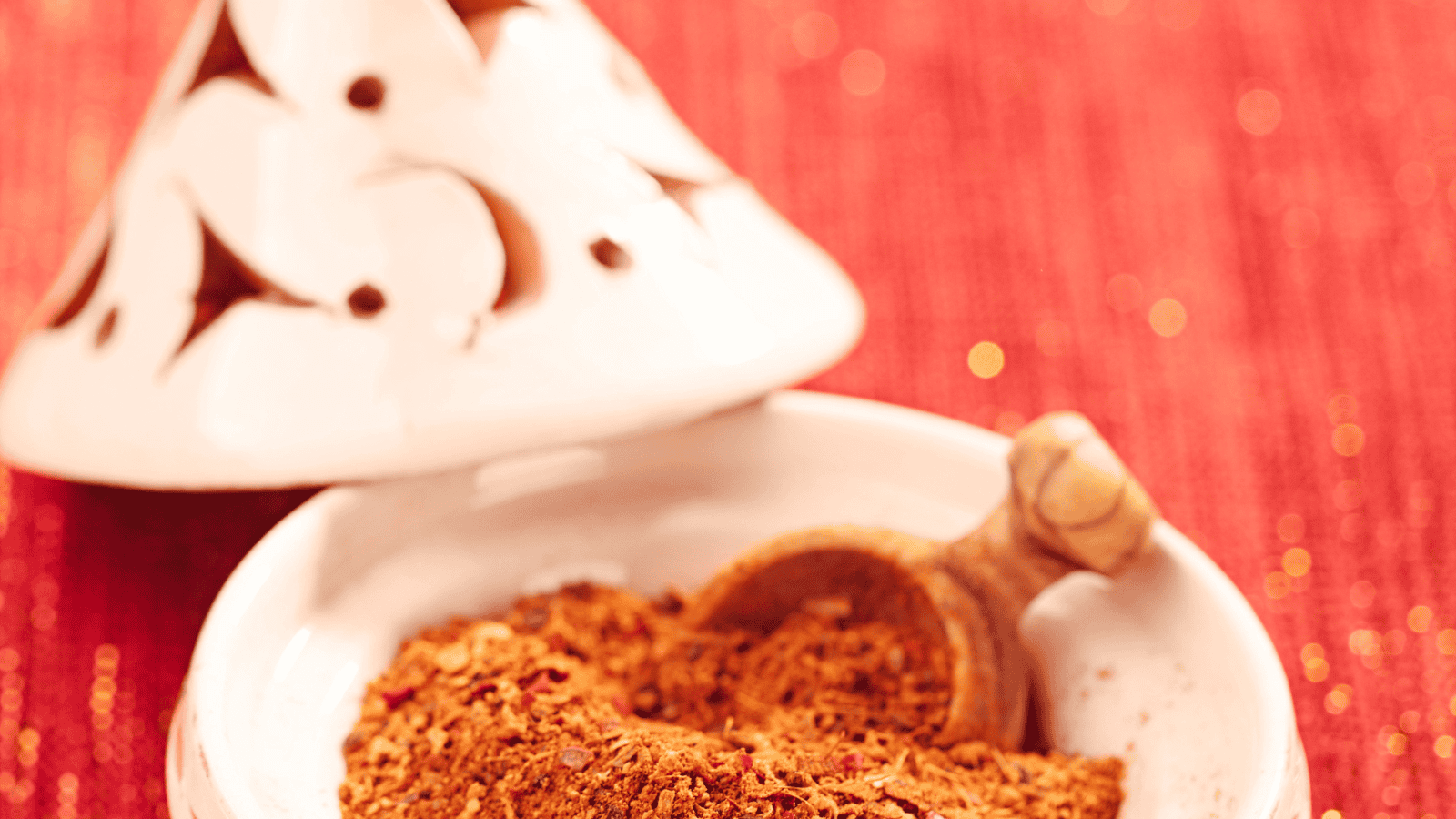A close-up of a wooden spoon in a white bowl filled with ground spices. The bowl is placed on a red textured surface, and a white ceramic jar with decorative cutouts is visible in the background.