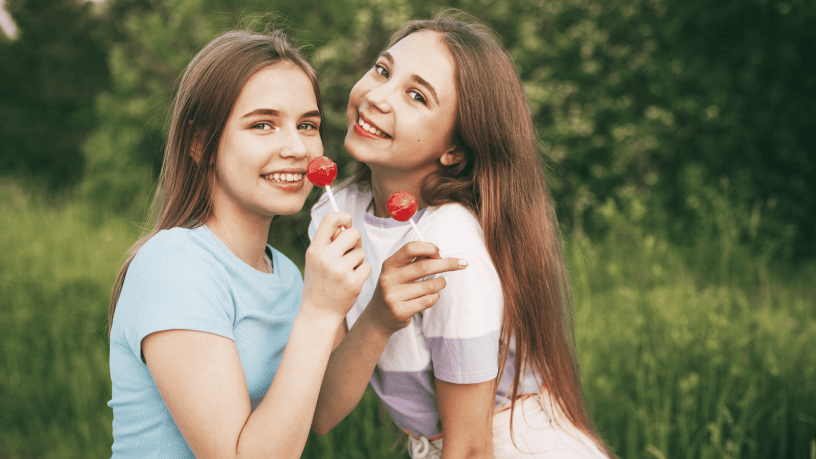 Two smiling young women stand close together outdoors, holding lollipops near their mouths. One wears a light blue shirt, and the other wears a white and purple shirt. They are in a grassy area with trees in the background.