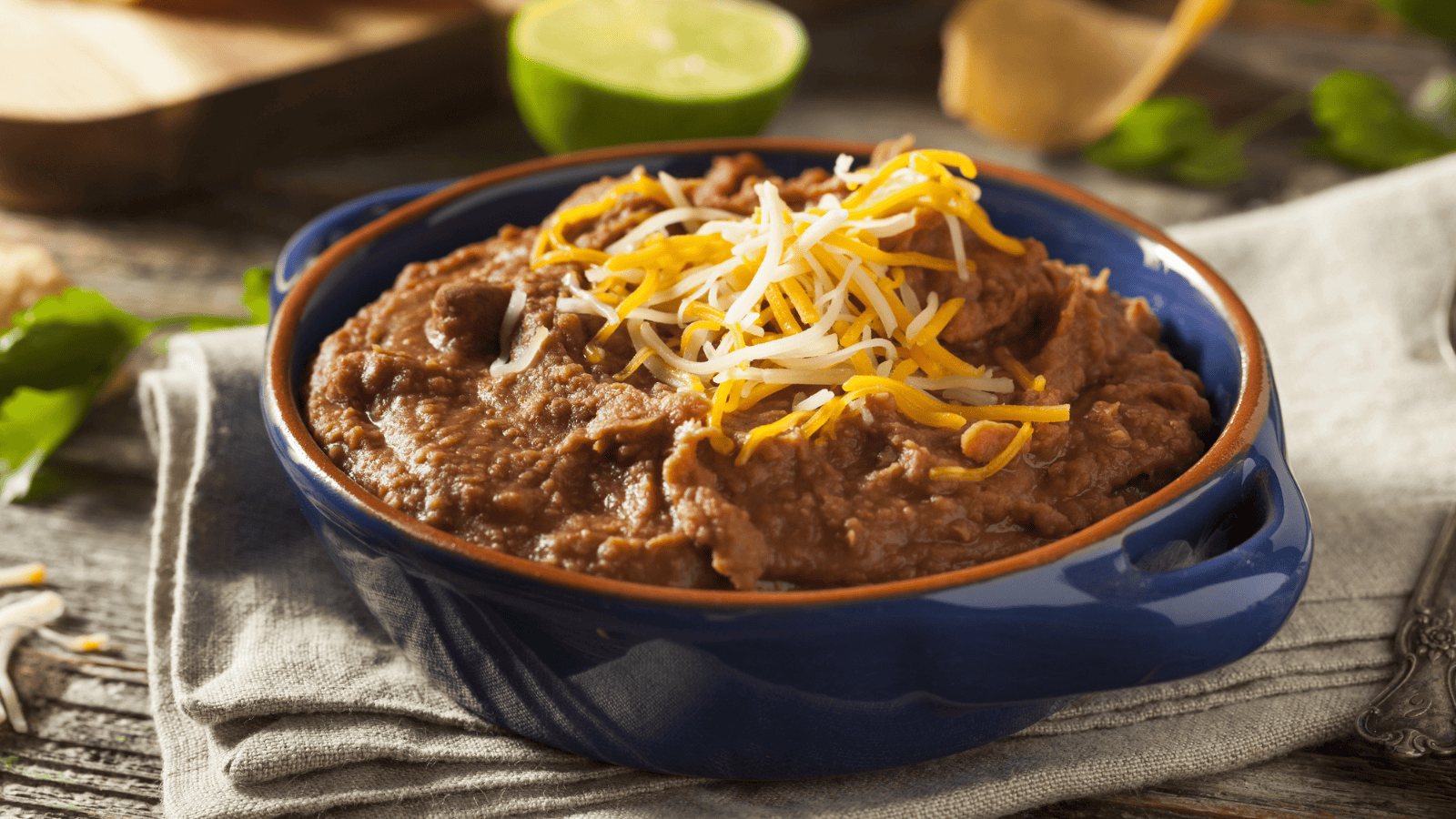 A blue ceramic bowl filled with refried beans, topped with shredded cheese. The bowl is placed on a beige napkin. In the background, there is a lime cut in half and scattered cilantro leaves.