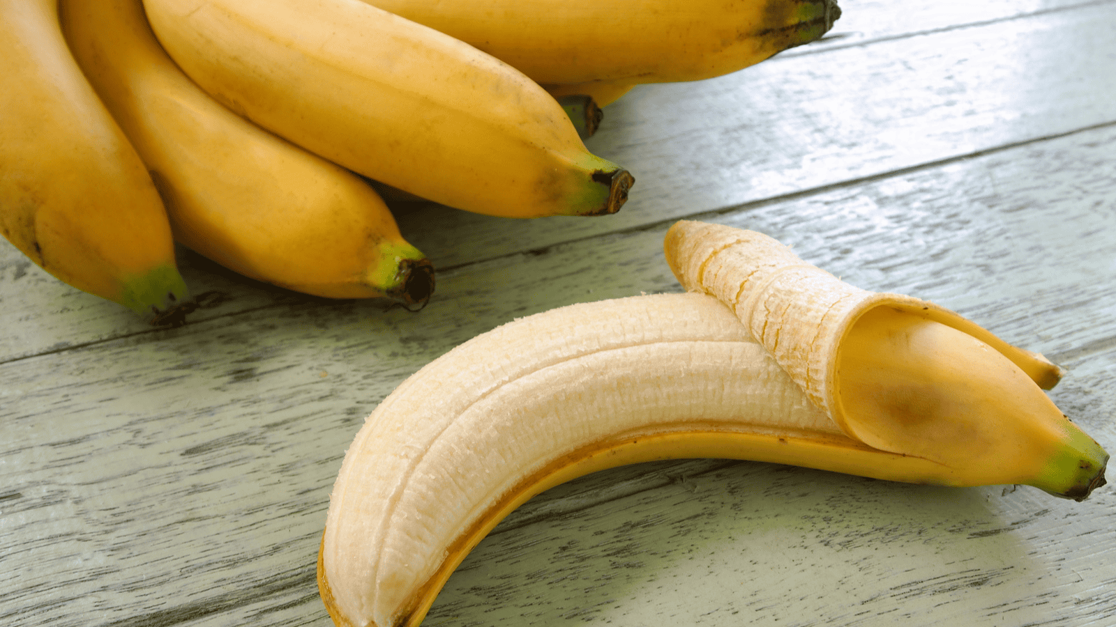 A bunch of bananas sits on a wooden surface. In the foreground, one banana is partially peeled, revealing the fruit inside. The wood has a weathered, light greenish-gray appearance.