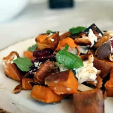 A white plate filled with roasted sweet potato cubes, goat cheese, beetroot, and fresh herbs showcases one of many non-boring salad recipes. A fork lies to the left of the plate, with a glass partially visible in the background at the top right.