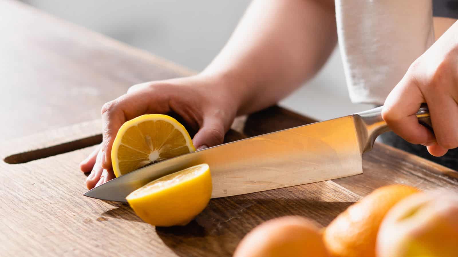 A person slicing a lemon in half with a large kitchen knife on a wooden cutting board, utilizing Grandma's kitchen secrets, with more whole citrus fruits visible nearby.