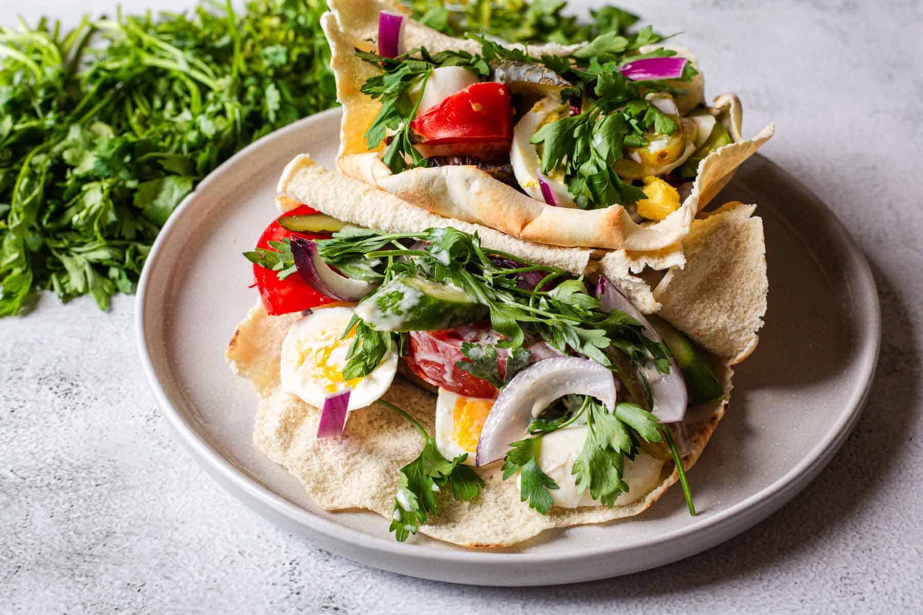 A plate with two pita sandwiches filled with sliced tomatoes, boiled eggs, red onions, fresh parsley, and cucumber, with more fresh parsley in the background.