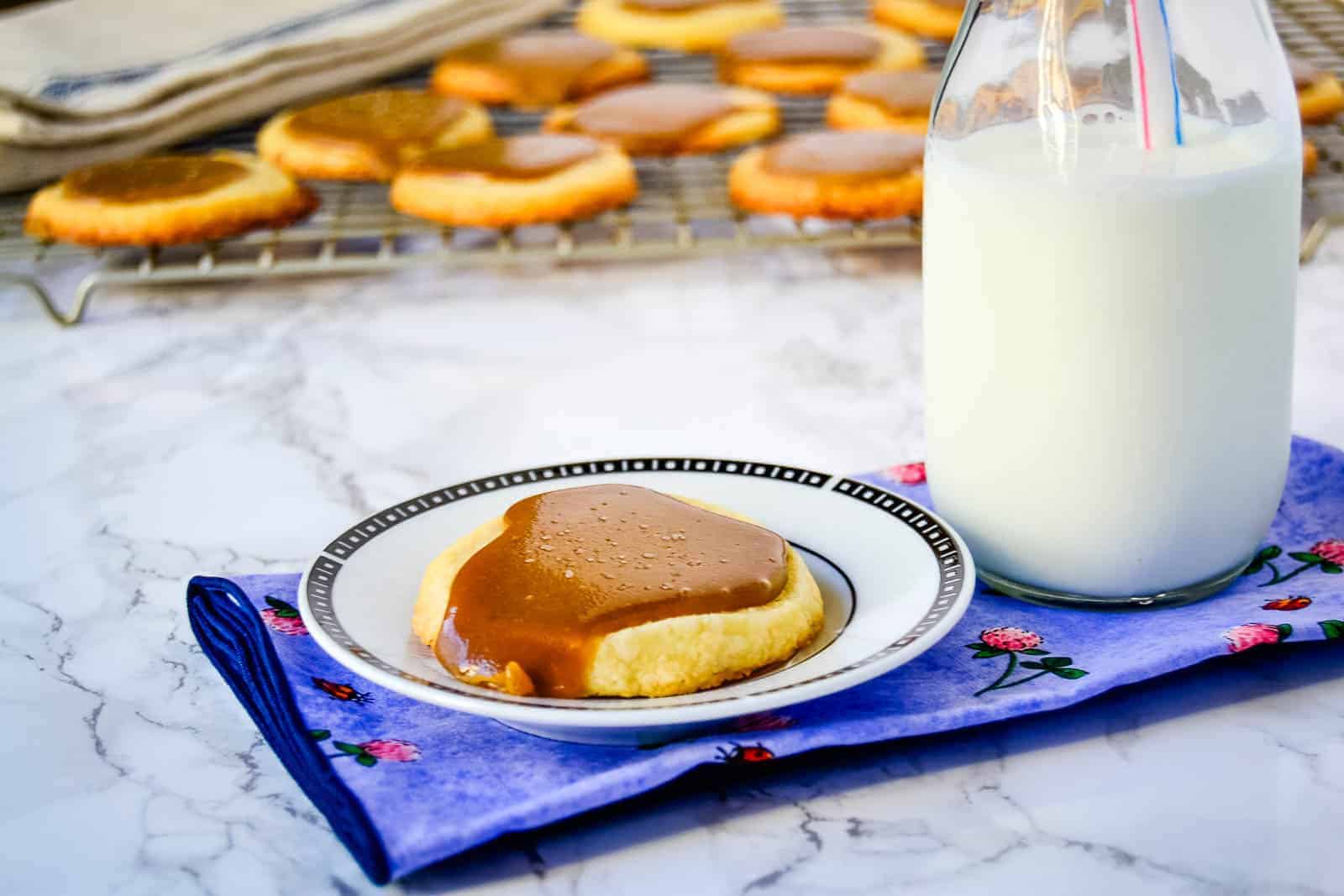 A plate with a caramel-topped pancake sits beside a glass of milk on a marble surface, with more pancakes on an auto draft cooling rack in the background.
