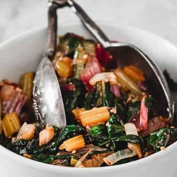 A white bowl contains a colorful salad made with chopped Swiss chard, including dark green leaves and pale to vibrant red stems. Slivers of onions are also visible. Two metal utensils rest in the bowl, ready for serving. The background is a light marble surface.