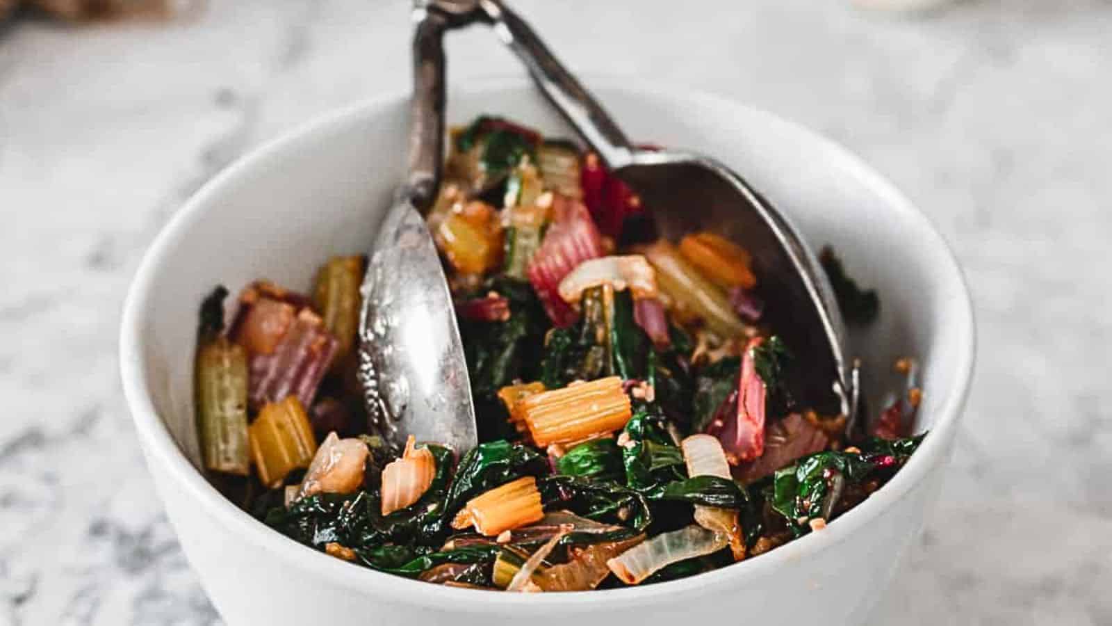 A white bowl contains a colorful salad made with chopped Swiss chard, including dark green leaves and pale to vibrant red stems. Slivers of onions are also visible. Two metal utensils rest in the bowl, ready for serving. The background is a light marble surface.