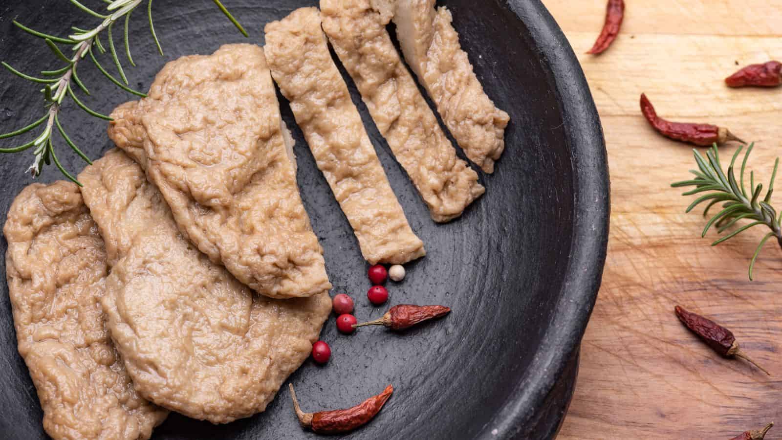 Slices of seitan arranged on a black plate, seasoned with rosemary, pink peppercorns, and dried chili peppers. Discover the meatless muscle power in this seitan with its textured, meat-like appearance.