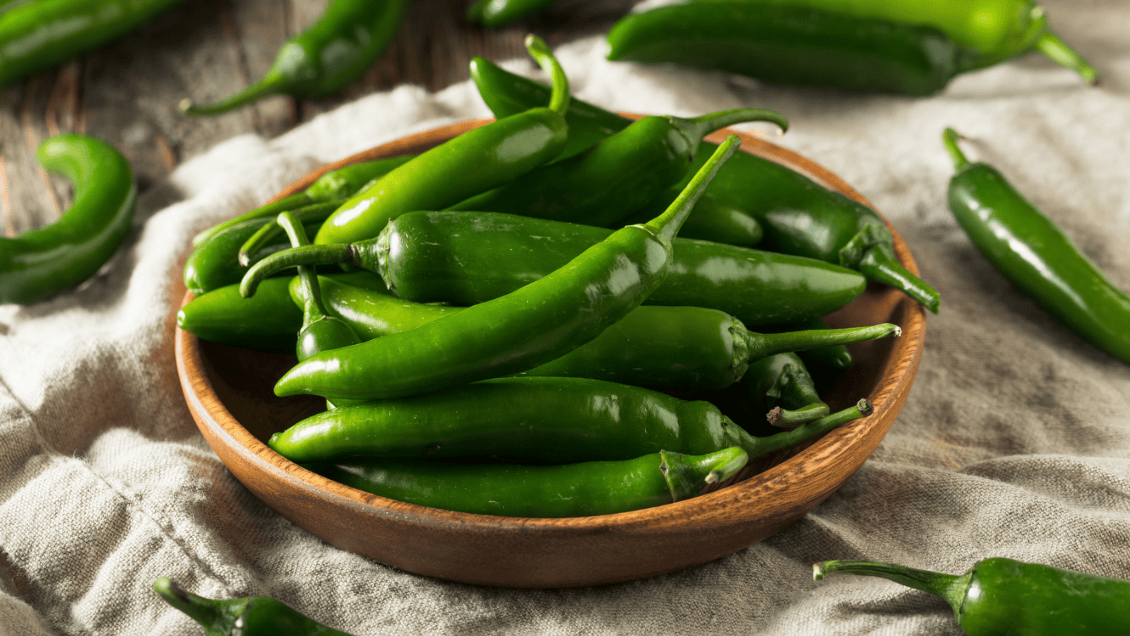 A wooden bowl filled with fresh green chili peppers sits on a cloth surface. Several additional chili peppers are scattered around the bowl. The peppers have a glossy, smooth texture and are elongated with pointed ends.
