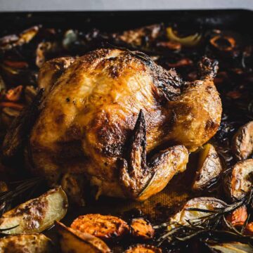 A roasted whole chicken sits on a baking tray surrounded by roasted vegetables, including carrot slices, onion pieces, and sprigs of rosemary. Two hands can be seen holding the edges of the tray.