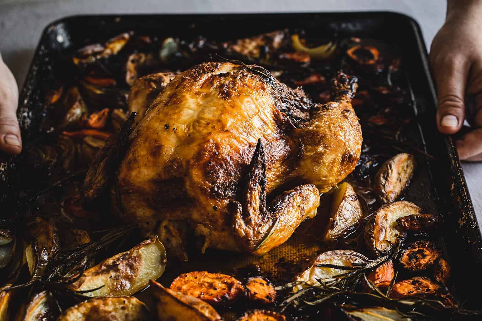 A roasted whole chicken sits on a baking tray surrounded by roasted vegetables, including carrot slices, onion pieces, and sprigs of rosemary. Two hands can be seen holding the edges of the tray.