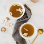 Two bowls of shiitake mushroom broth with sliced mushrooms and green onions, on a white marble surface with a draped gray cloth and a golden spoon to the side.