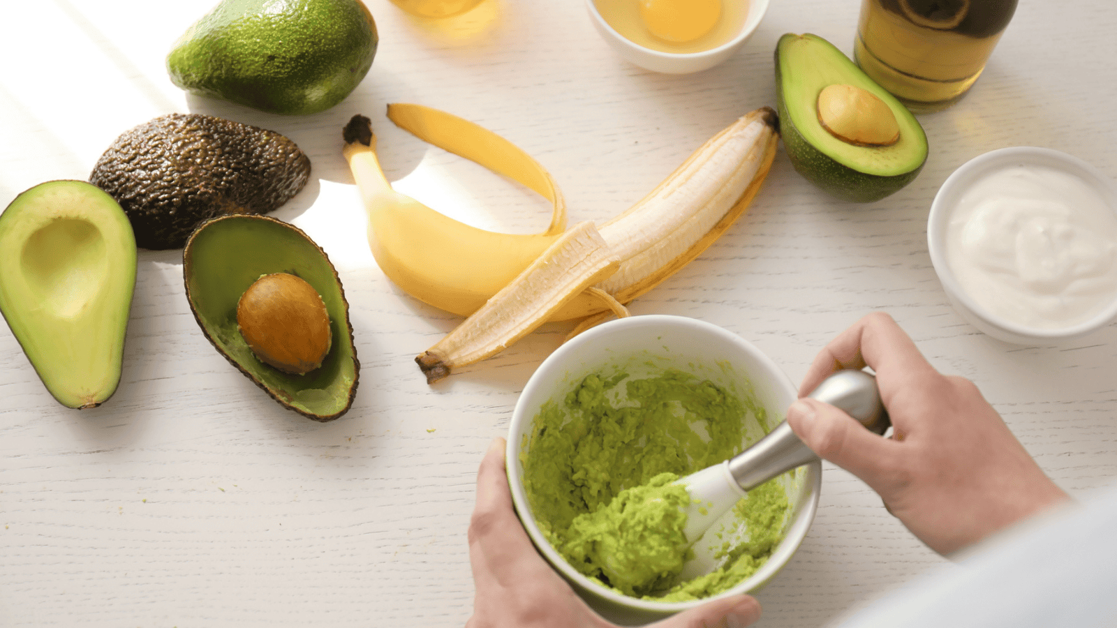 A person mashes avocado in a bowl. The table has whole and halved avocados, a peeled banana, a bowl of white yogurt, and a bottle of oil. There are remnants of another avocado and a banana peel in the background.