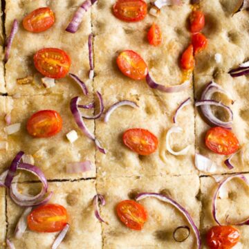 A rectangular focaccia bread is shown, topped with cherry tomato halves and thinly sliced red onions. This delightful breakfast bread is cut into square pieces and features a light golden-brown crust. The surface is dotted with dimples, adding to its rustic charm.