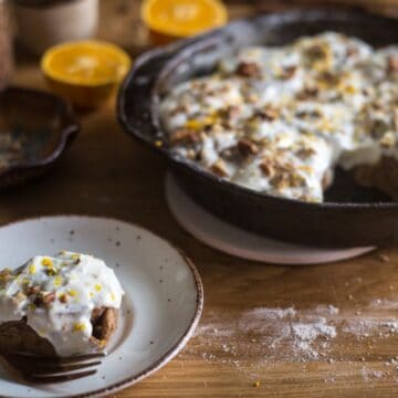 A skillet of freshly baked cinnamon rolls topped with white icing and sprinkled with orange zest, alongside a plate with a single served roll and a fork on a wooden table, perfect for celebrating Mother's Day.