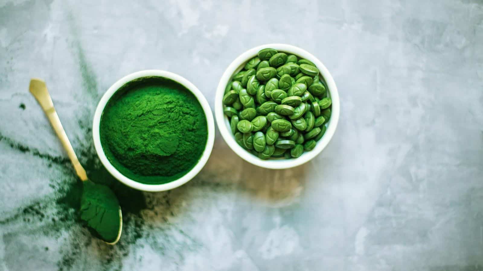 Two bowls on a gray surface; one contains veggie-powered proteins in the form of green powder with a spoon, the other filled with green capsules. The setup suggests a focus on nutritional or health supplements.