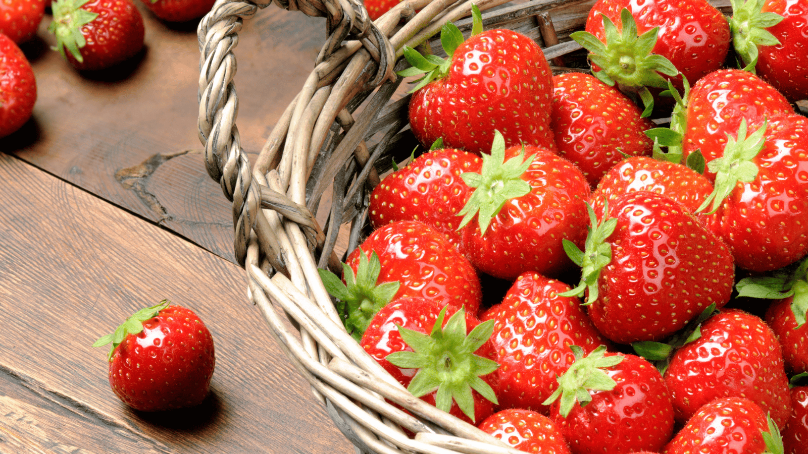 A close-up of a wicker basket filled with fresh, red strawberries with green leaves. The basket is on a wooden surface, and a few strawberries are scattered around outside the basket.