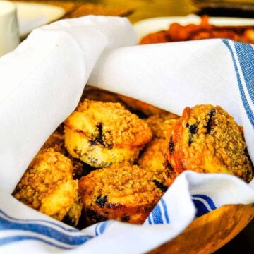 A wooden bowl lined with a white and blue striped cloth contains several streusel-topped breakfast breads. A white cup and other dishes are partially visible in the background on a table.