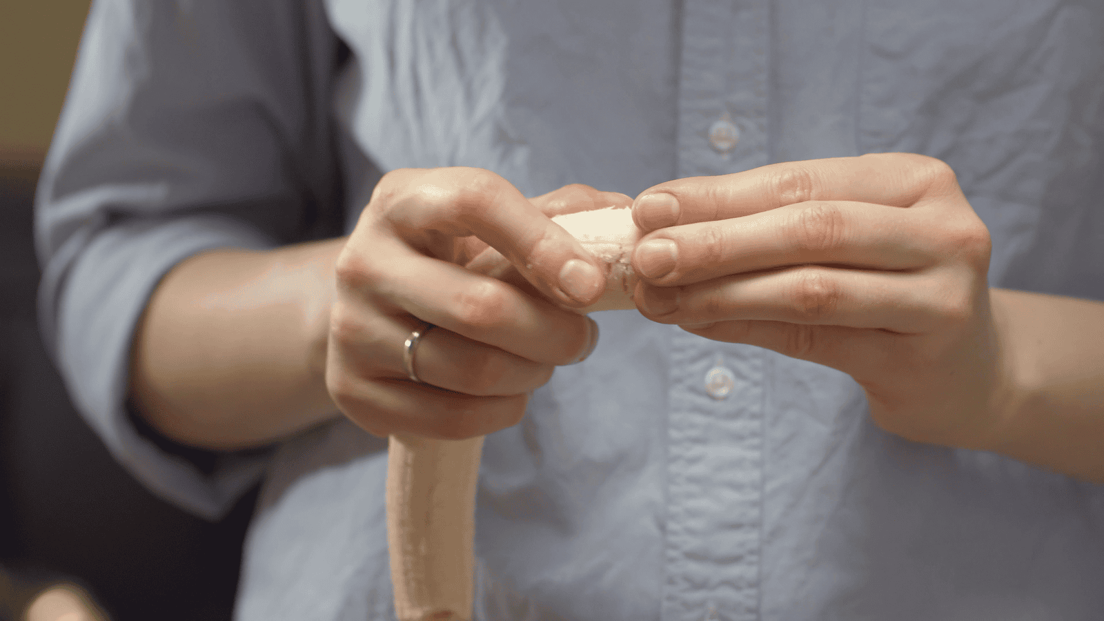 A person wearing a light blue button-up shirt is peeling a banana. Their hands are holding the top part of the banana as they remove the peel. The person is wearing a gold ring on the ring finger of their left hand.