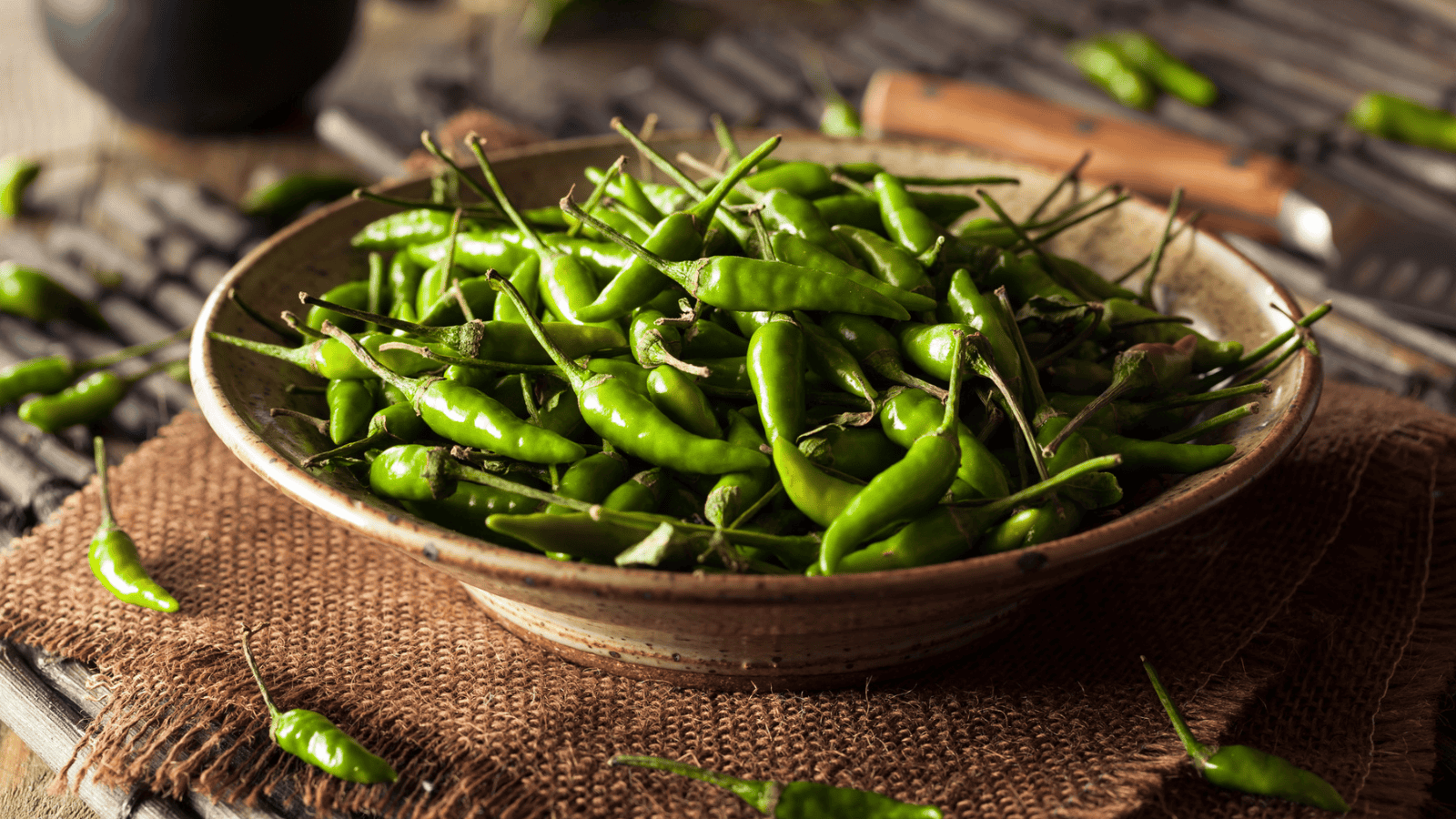 A bowl filled with numerous green chili peppers sits on a rustic woven mat. Some chili peppers are scattered around the bowl, and a knife is partially visible in the background. The setting appears to be a wooden surface.