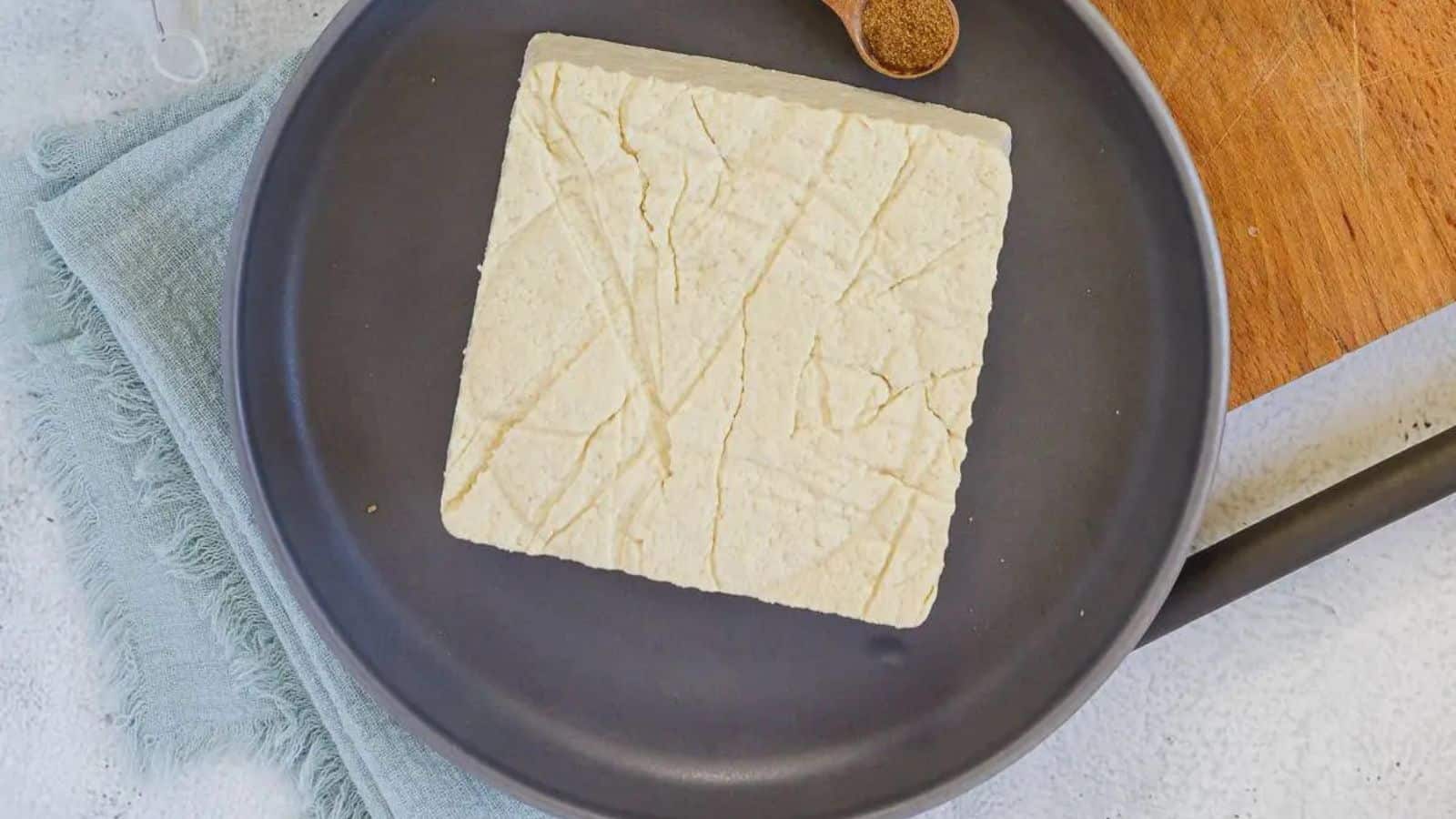 A block of tofu, rich in veggie-powered proteins, is centered on a dark gray plate, with a wooden cutting board and a spoon of spices nearby. The setting has a light, textured background.