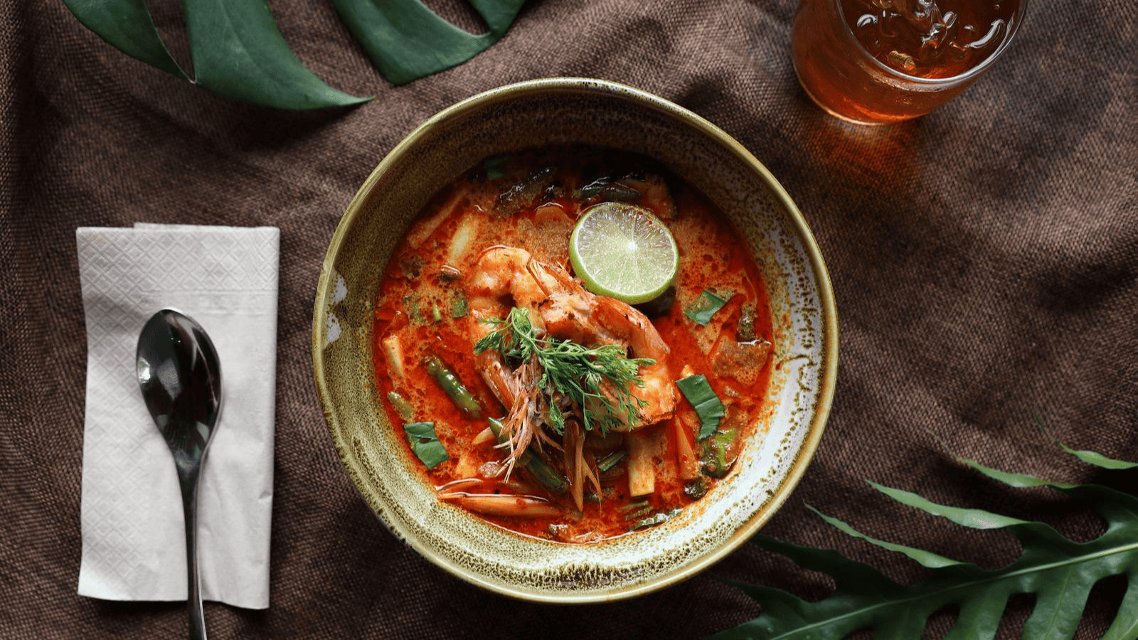 A bowl of spicy red shrimp soup garnished with a lime wedge, herbs, and sliced vegetables. The bowl is on a brown cloth, with a napkin and spoon to the left. A glass of iced tea is to the top right of the bowl. Leaves decorate the background.