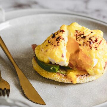 A plate with eggs benedict featuring a poached egg, hollandaise sauce, and avocado slices on an english muffin, garnished with spices, accompanied by gold cutlery. Perfect for Mother's Day recipes.