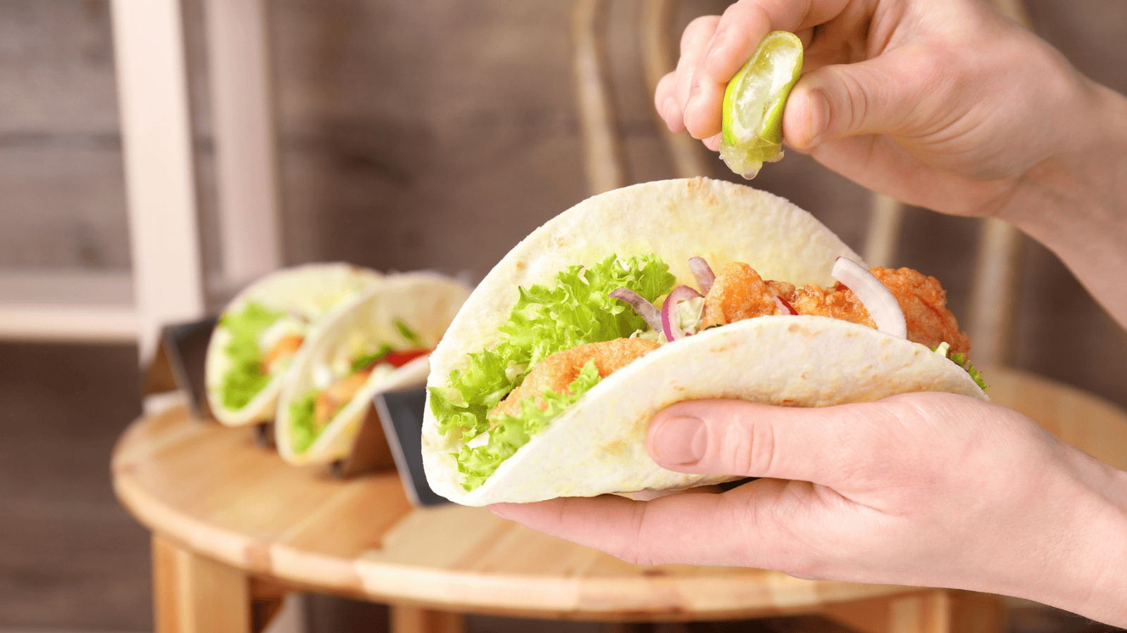 A person squeezes a lime wedge over a taco filled with fried chicken, lettuce, and red onion. Two other tacos are positioned on a wooden table in the background, held upright in a metal taco holder.