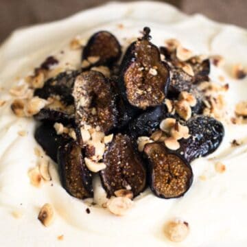 A retro close-up of a dessert featuring a layer of cream topped with sliced figs and chopped nuts. The creamy texture and dark figs provide a striking contrast in color and texture. The background appears to be a neutral-toned cloth, reminiscent of classic dessert recipes.