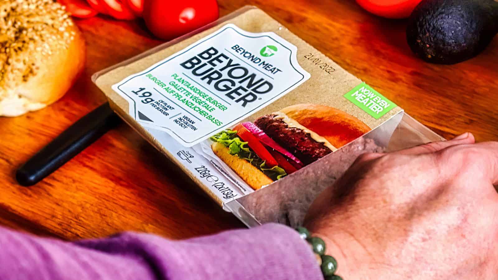 A person's hand reaches for a Beyond Burger package on a wooden kitchen counter, surrounded by fresh vegetables like tomatoes and avocados. The package highlights the burger as plant-based and high in Veggie-Powered Proteins.
