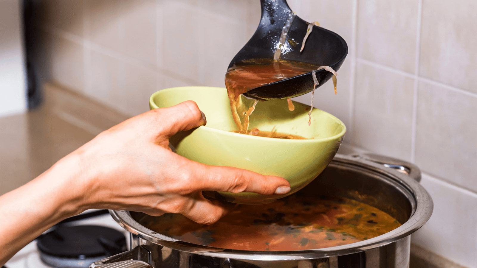 A person is serving soup from a pot into a light green bowl using a black ladle. The soup appears to be broth-based with visible ingredients like vegetables. The scene is set in a kitchen, with the pot on a stovetop and a tiled backsplash visible.